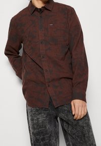 Röd och svart mönstrad långärmad skjorta med knäppning framtill, spetsig krage och bröstficka, tillsammans med texturerade mörkgrå byxor.