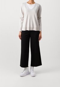 Jasnoszara, długorękawowa, swetrowa bluzka z dekoltem w serek, nałożona na białą koszulę, połączona z czarnymi, szerokimi spodniami i białymi sneakersami za kostkę.