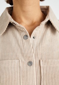 Camicia in corduroy beige con colletto, caratterizzata da righe verticali, bottoni a pressione in metallo e due tasche sul petto. Texture liscia e vestibilità sartoriale.