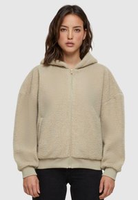 Giacca beige con cappuccio e zip realizzata in morbido fleece testurizzato, con maniche oversize, polsini a coste e due tasche laterali.