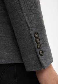 Szary rękaw swetra dzianinowego z czterema czarnymi guzikami wzdłuż mankietu. Tekstura jest miękka, o gładkim, równomiernym splocie.