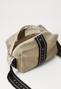 Borsa a rete beige con chiusura a zip, dotata di cinturini neri con marchio. Il design è compatto e rettangolare con una superficie texturizzata.