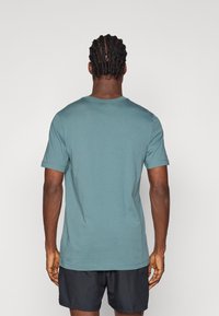 Τυρκουάζ βαμβακερό μπλουζάκι με κοντά μανίκια, στρογγυλή λαιμόκοψη και χαλαρή εφαρμογή, προφανές από την πλάτη. Λείος υφασμάτινος συνδυασμός, χωρίς ορατά σχέδια ή διακοσμητικά.