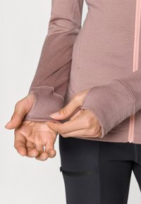 Casaco desportivo rosa claro com buracos para os polegares nas mangas, fecho de zipper e textura suave, combinado com leggings pretas ajustadas.