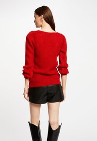 Maglione rosso lavorato a maglia con una finitura testurizzata, maniche a sbuffo e scollo ampio, abbinato a pantaloni corti in pelle nera e stivali neri alti.
