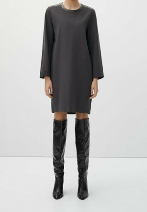 Femme portant une robe ample gris foncé à manches longues, longueur genou, associée à des bottes en cuir noires montant jusqu’au genou, debout devant un fond uni.
