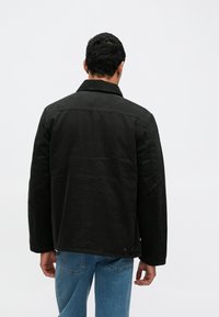 Czarna bawełniana kurtka z klasycznym kołnierzem, detalami na guziki i prostym krojem. Noszona z jasnoniebieskimi dżinsami, widziana od tyłu.