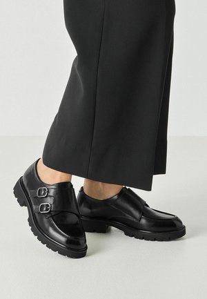 Schwarze Lederschuhe mit doppelter Schnalle und klobiger Sohle, getragen zu schwarzen, kurz geschnittenen, weiten Hosen auf hellem Hintergrund.