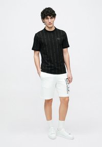 Czarny T-shirt z krótkim rękawem z powtarzającym się wzorem logo, zestawiony z białymi szortami z pionowym nadrukiem nazwy marki. Całość dopełniają białe trampki.