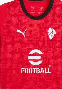 Rød fodboldtrøje lavet af tekstureret stof. Har hvidt Puma-logo, løvegrafik og "FOOTBALL" tekst i fed skrift. Sort kraveaccent.