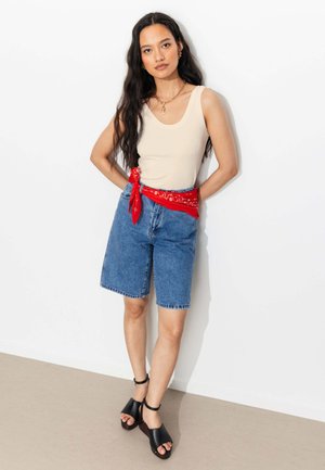 Femme aux longs cheveux foncés portant un débardeur beige, un short en jean longueur genoux, une ceinture bandana rouge et des talons ouverts noirs, debout contre un mur blanc.