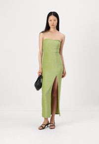 Vestido maxi verde, sem alças, ajustado, com uma fenda lateral, feito de um tecido brilhante. Modelo a segurar uma mala preta, usando sandálias pretas com tiras.