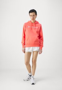 Felpa corallo con tasca frontale e cappuccio con lacci. Presenta un grande logo "Champion" bianco. Abbinata a pantaloni corti bianchi e scarpe da ginnastica.