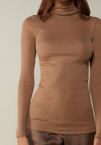 Femme portant un pull à col roulé ajusté, côtelé, à manches longues de couleur marron, et un pantalon marron foncé, se tenant devant un fond uni.
