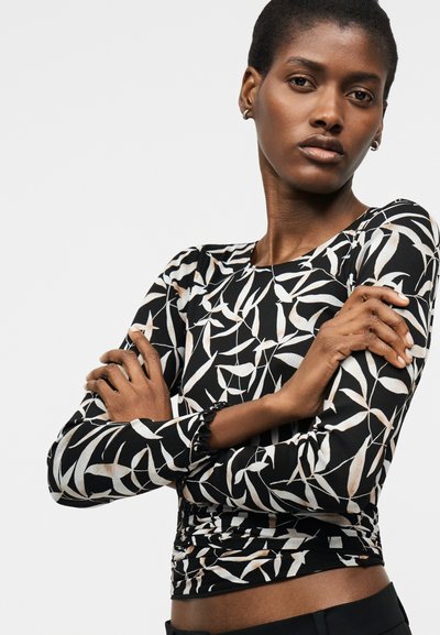 Γυναίκα με κοντά μαλλιά που φοράει μακρυμάνικη μπλούζα με ασπρόμαυρο σχέδιο, με σταυρωμένα τα χέρια, μπροστά σε απλό φωτεινό φόντο.