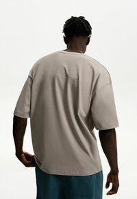 Tricou bej oversize cu mâneci scurte, cu detaliu cusătură pe spate, realizat dintr-un material moale din bumbac, purtat cu pantaloni scurți teal.