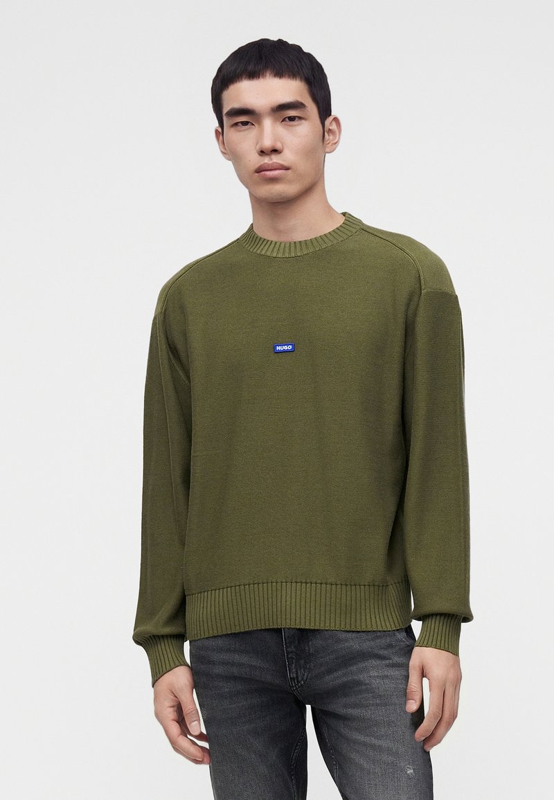 Jeune homme portant un pull vert olive avec un petit logo bleu sur la poitrine et un jean gris foncé, debout devant un fond clair uni.