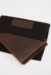 Brown leather wallet inside a black fabric pouch with a branded leather tag. Features visible stitching and smooth texture.