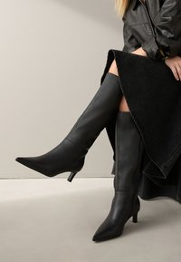 Personne portant des bottes en cuir noir montantes jusqu'aux genoux, à bouts pointus et talons kitten, assorties à une jupe et une veste noires.