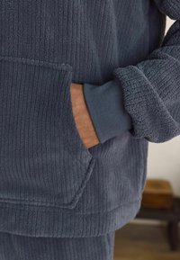 Dark grey fleece hoodie with vertical ribbed texture, featuring a front pocket and ribbed cuffs. Soft fabric is visible and tactile.