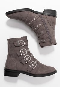 Botas de ante gris hasta el tobillo con múltiples hebillas plateadas, cierre de cremallera y un tacón bajo negro. Punta redondeada y diseño estructurado.
