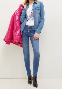 Mujer vestida con una camisa de mezclilla azul y jeans, sosteniendo una chaqueta acolchada de color rosa brillante sobre el hombro, de pie con botas de tacón negro.