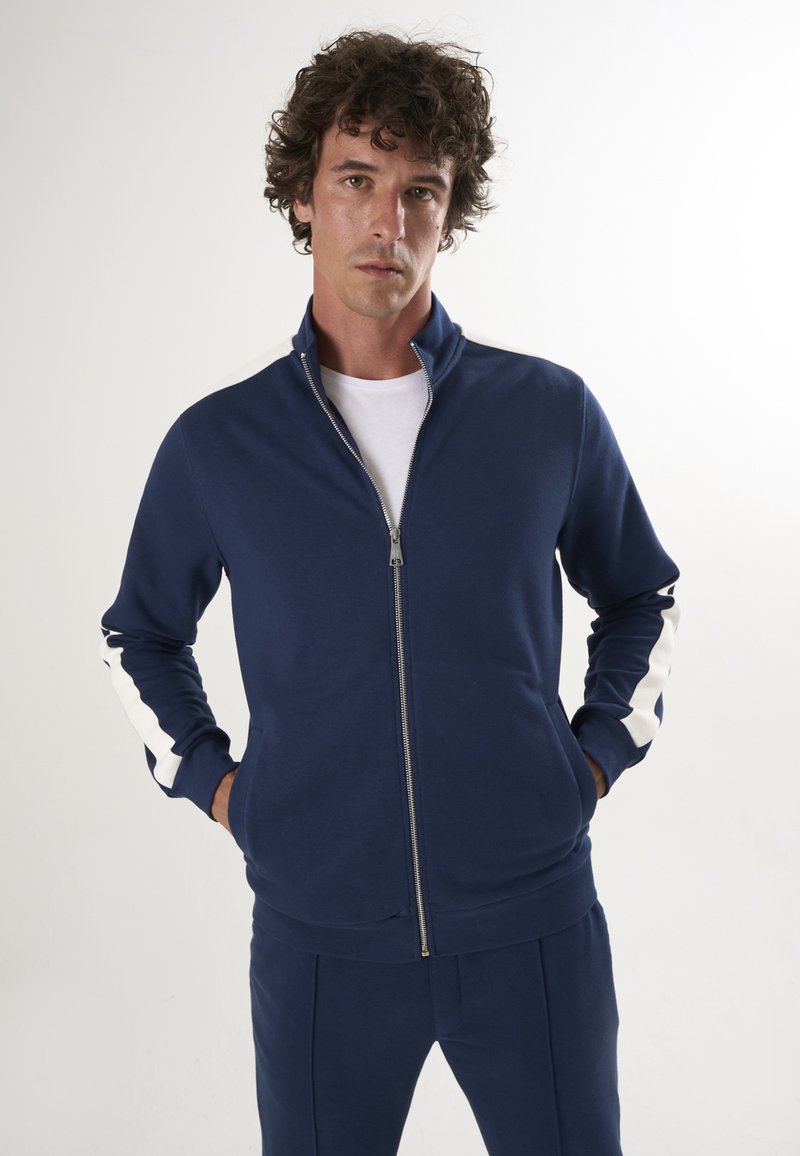 Marineblå lynlåset sportsjakke lavet af blødt stof, med hvide sidestriber og en opretstående krave. Stylet med en hvid t-shirt under.