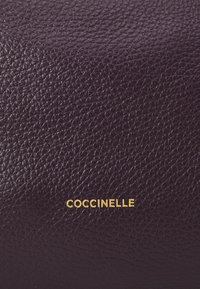 Portafoglio in pelle viola scuro con una finitura texturizzata. Caratterizzato dal logo dorato "Coccinelle" in basso. Design compatto e elegante.