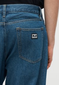 Πίσω όψη ατόμου που φοράει μπλε τζιν με τετράγωνο ασπρόμαυρο λογότυπο στο πίσω τσεπάκι, με εμφανές το δεξί χέρι.