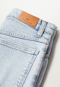Jean en denim bleu clair plié avec une étiquette en cuir marron portant l'inscription « MNG Authentic Denim » au-dessus de la poche arrière.