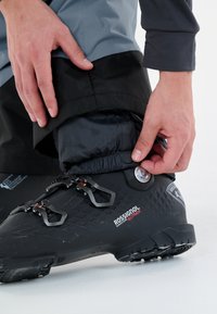 Svart skidboot med texturerad yta, spännen och en cirkulär justeringsknapp nära vristen, tillsammans med justerbara lagerbyxor.