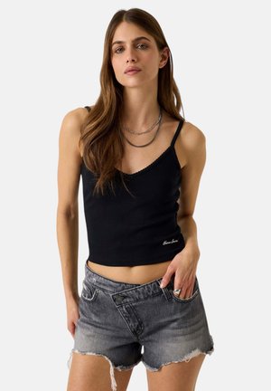 Giovane donna con lunghi capelli castani che indossa un top nero corto e pantaloncini di jeans grigi a vita alta con bordi sfrangiati.