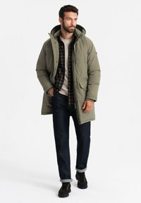 Olivgrüne gep padded Jacke mit Kapuze, mit großen Fronttaschen. Getragen über einem karierten Hemd, kombiniert mit dunklen Jeans und schwarzen Stiefeln.