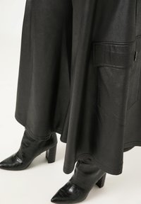 Schwarzes Kunstlederkleid mit einer strukturierten Oberfläche, abgerundetem Saum und einer seitlichen Tasche. Kombiniert mit schwarzen spitzen Absatzstiefeln.