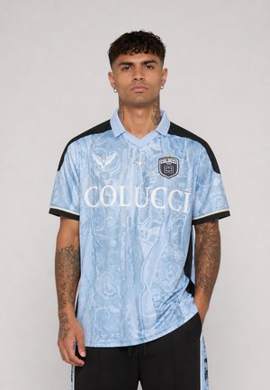 Hombre de pie que lleva una camiseta deportiva Colucci azul claro con paneles negros en los hombros y diseño estampado, manos relajadas a los lados.
