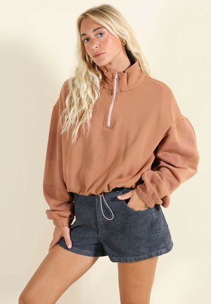 Femme aux longs cheveux blonds portant un pull marron à demi-fermeture éclair et un short taille haute à motif sombre, posant avec une main dans la poche.