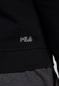 Sudadera negra con textura acanalada y un logo en blanco y rojo en el lado izquierdo. Presenta un color sólido y un diseño sencillo.