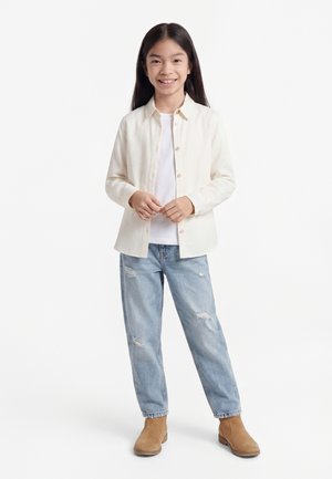 Hosszú sötét hajú, mosolygó lány, fehér, gombos ingben, fehér trikó felett, szakadt kék farmerral és bézs bokacsizmában.