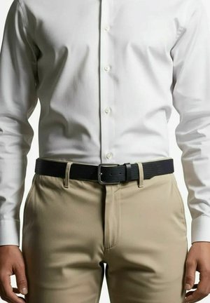 Man die beige broek, zwarte riem en wit overhemd met knopen draagt, staand met ontspannen armen langs het lichaam tegen een effen achtergrond.