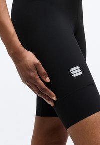 Κοντινό πλάνο ενός χεριού που ακουμπάει σε μηρό με μαύρο ποδηλατικό σορτς που φέρει λευκό λογότυπο στο πόδι.