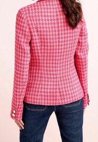 Sako s červenobílým vzorem houndstooth, pevné střihy, výrazné knoflíky a dlouhé rukávy. Kombinováno s tmavými džínovými kalhotami.