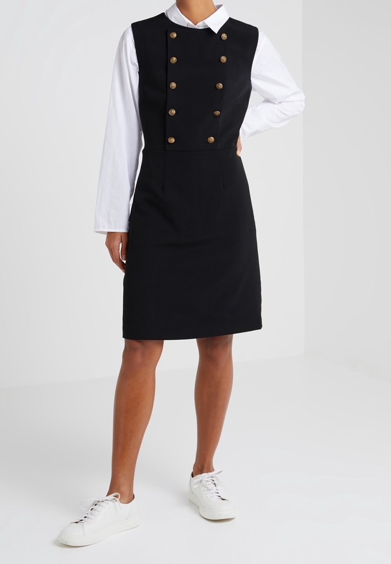 Musta A-linjainen mekko ilman hihoja, jossa on kultaisia nappiyksityiskohtia ja alla valkoinen pitkähiha-paita. Yhdistetty valkoisiin lenkkareihin.