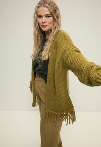 Olivgrüner Strickcardigan mit grober Textur und fransigen Kanten, getragen über einem schwarzen Grafik-T-Shirt und hellbraunen Hosen.