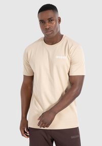 Beige t-shirt met korte mouwen van katoen, met een ronde halslijn en een klein wit logo op de linkerborst. Soepele textuur.