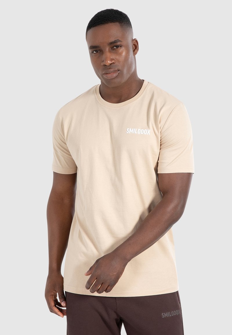 Beige t-shirt met korte mouwen van katoen, met een ronde halslijn en een klein wit logo op de linkerborst. Soepele textuur.