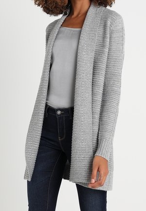 Femme portant un cardigan à côtes gris clair ouvert sur le devant, par-dessus un caraco gris et un jean bleu foncé, debout devant un fond uni.