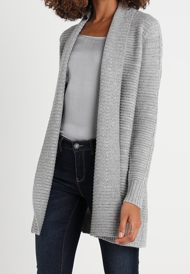 Femme portant un cardigan à côtes gris clair ouvert sur le devant, par-dessus un caraco gris et un jean bleu foncé, debout devant un fond uni.
