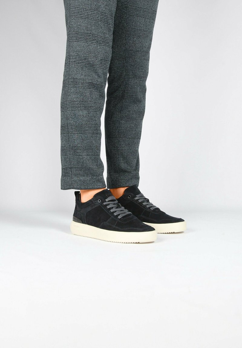 Personne portant un pantalon gris à carreaux et des baskets basses noires avec des semelles blanches, debout sur une surface blanche.