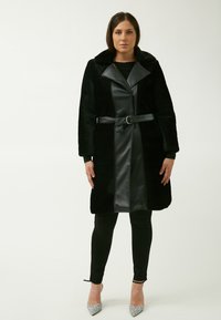 Cappotto nero con una texture di pelliccia e parte anteriore in pelle, caratterizzato da un ampio colletto, vita con cintura e maniche lunghe. Indossato sopra leggings neri e tacchi.