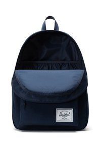Navy blue backpack with a flap, textured material, and a front pocket. Features a branded patch and a top carry handle. Interior pocket included.